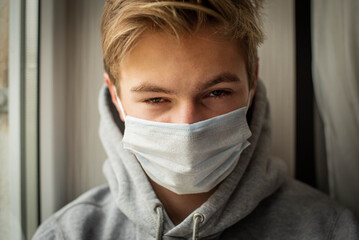 Sad young ill boy looking sad with protective mask at home behind window in quarantine and lockdown missing school and freedom during Covid-19 Coronavirus worldwide pandemic