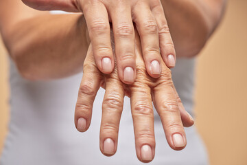 Cropped photo of a mature woman rubbing moisturizer on hands while standing against beige background