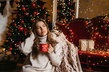 Young woman smiling, relaxing at home, enjoying Christmas. Beautiful girl drinking tea at home, sitting near the Christmas tree