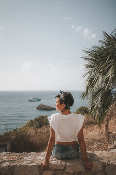 Beautiful woman with short hair looking at the sea smiling while sitting down over a cliff in Ibiza wearing denim shorts. Rear view