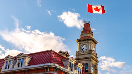 Victoria, British Columbia, Canada Victorian-style building with a huge Canadian flag on a post waving in the wind against a blue sky and fluffy clouds. - obrazy, fototapety, plakaty