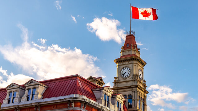 Victoria, British Columbia, Canada Victorian-style Building With A Huge Canadian Flag On A Post Waving In The Wind Against A Blue Sky And Fluffy Clouds.