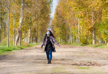 new normal Autumn walk outdoors - young happy and pretty Asian Korean woman in face mask walking cheerful at beautiful city park in vibrant yellow and orange tree leaves