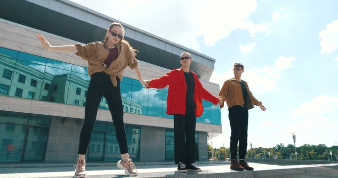 Three stylish teenagers stand together outdoors against near glass blue building, wear trendy bright shirts and sunglasses, hold hands, make wave with their arms and shoulders, perform dance element