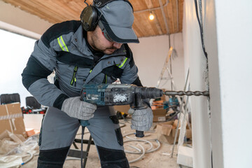 a worker, dressed in overalls, glasses and special headphones for noise reduction, drills a hole in the wall for wires