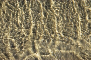 Fototapeta na wymiar Ripples on the water, sea water near the coast, sand is visible through the water, top view