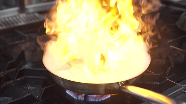 Skilled chef closes frying pan with delicious mussels and scallops at bright burning flame with metal lid on contemporary stove in restaurant kitchen extreme close view.