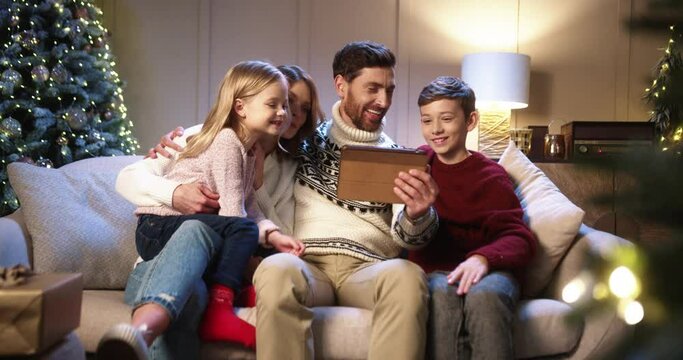 Caucasian cheerful family with children sitting in decorated room with christmas tree and videochatting on tablet Parents with kids speaking on video call on New Year's Eve. Holidays concept
