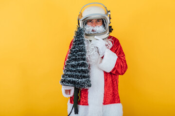 Fototapeta na wymiar Santa Claus with astronaut helmet holding a Christmas tree on yellow background. Christmas concept