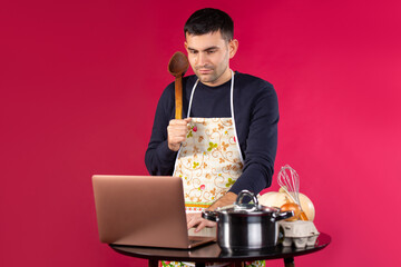 A serious man in the kitchen in an apron and with a spatula. Looking for a new recipe on a laptop....