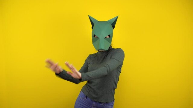 Young woman in a cardboard jackal mask dances on a yellow background. Woman in a green sweater and mask.