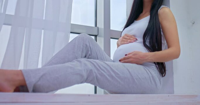 Beautiful pregnant woman sits and touches her tummy, feels the movement of the baby. Happy woman enjoying her pregnancy, sitting by the window and smiling. 4k, ProRes