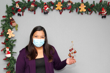 Latin woman with protection mask, santa claus hat and christmas arvol with spheres in christmas...