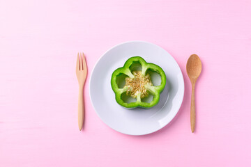 Sliced green bell pepper on white plate with wooden fork and spoon on pink background, Top view