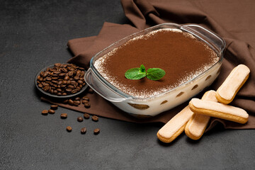 Traditional Italian Tiramisu dessert in glass baking dish and savoiardi cookies on concrete background
