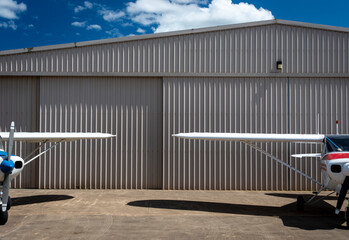 two aircraft, partially visible, white and blue and white and red, against a grey hanger and blue sky with clouds © Randal