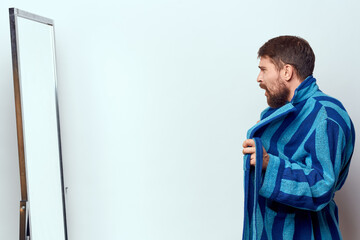 a man in a blue robe examines himself in a mirror in a bright room cropped view