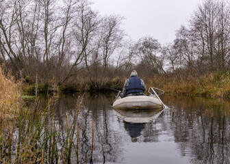 gray and cloudy day, fisherman in a white boat, river bank with bare trees and bushes, shore reflection in river water