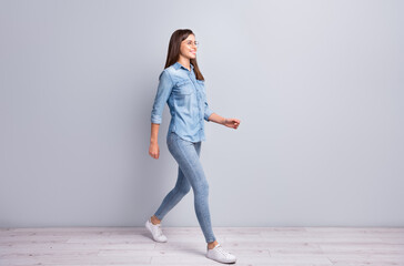 Full size profile photo of optimistic sweet girl walking wear shirt jeans sneakers isolated on concrete grey wall background