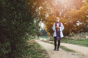 Young priest in a medical mask is standing on a country road
