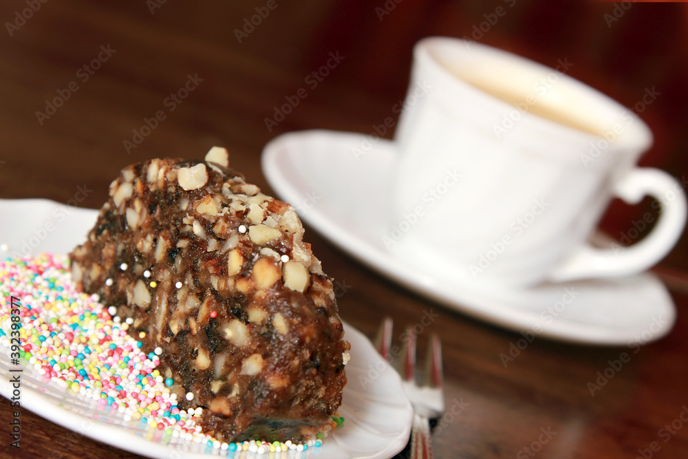 Poster focus shot of a piece of cake with walnuts and sprinkles with a cup of hot coffee in the background