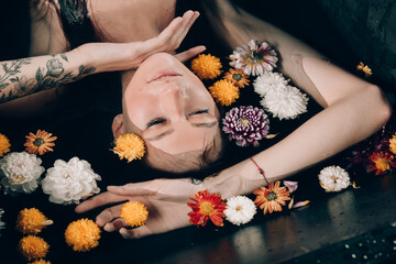 Young pretty brunette woman in bath with water and flowers. Naked model during an unusual photo shoot in a black bath