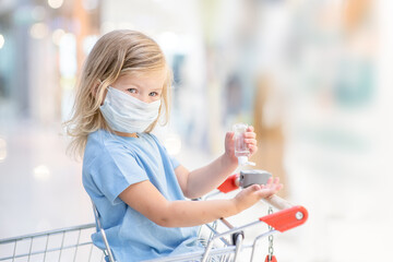 Little girl wearing protective facemask during coronavirus and flu outbreak sits inside shopping cart and applies sanitizer for cleaning hands in public crowded place. Empty space for text