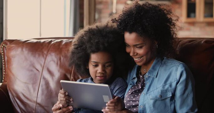 Little girl and mother sit on couch with tablet device. African ethnicity family spend time on internet, enjoy educational web sites for kid development, modern tech usage for fun or education concept