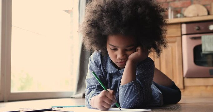 Cute african girl with curly hair lying on wooden floor with under floor heating system, little kid draw with pencil in sketchbook in cozy domestic kitchen. Hobby, free time, modern warm house concept