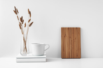 A mug on a stack of books and a transparent vase of dried flowers, a wooden board against the wall. Eco-friendly materials in interior decor. Copy space, mock up.