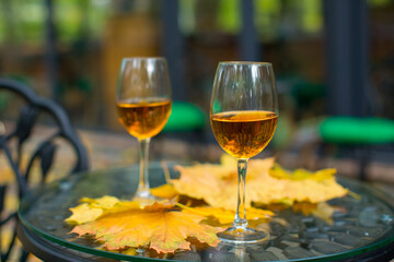 Two glasses of cognac-colored white wine on a table covered with yellow leaves. Two chairs with green upholstery. The sidewalk strewn with yellow leaves on the background of the restaurant.