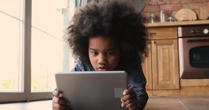 Little african girl lying on warm floor with tablet, look at device screen watch cartoons, listen audio book, close up. Addiction of modern tech usage, parental control, safety, gadget overuse concept
