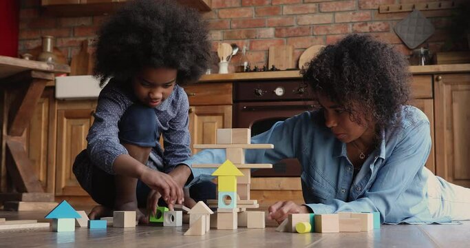 Happy african mother play wooden blocks with little daughter on warm kitchen floor, family having fun stacking cubes building tower spend free time at modern home. Leisure together with kids concept