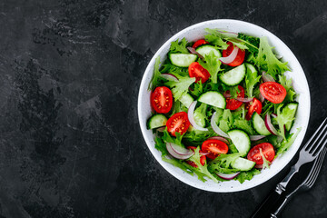 Healthy green salad with fresh tomato, cucumber, red onion and lettuce in bowl on dark stone background.