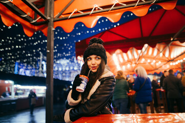 Obraz na płótnie Canvas Cute girl in warm clothes drinks a warming drink from a cup on the background of the Christmas market in the evening, decorated with light. Lady spends time in the evening at the fair.
