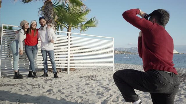 Two young heterosexual couples in sea winter vacation posing for a picture - Caucasian man kneeling taking a photo to his thumbs up friends on a beach of a vacation resort in Golfo Aranci, Sardinia