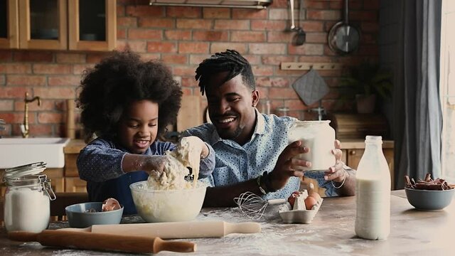 African dad with dreadlocks pour flour while curly haired daughter knead dough in bowl, family prepare surprise sit at dining table in kitchen. Spend time together develop kid, pleasant chores concept