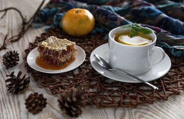 Breakfast with tea and lemon. White tea Cup on a brown woven napkin with honeycombs of buckwheat honey. Fir cones. A tangerine, a branch of lemongrass.