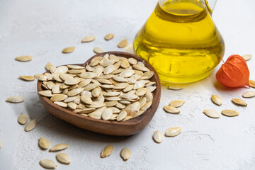 Fried pumpkin seeds lies in heart shaped wooden bowl on the gray background next to glass bottle of pumpkin seed oil and orang physalis flower. Natural organic food theme.