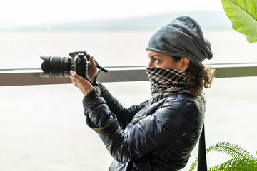 Young female photographer with bandana standing near large window and taking picture of river