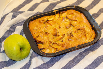 Apple charlotte in the black cake tin and a green apple on the tablecloth. 