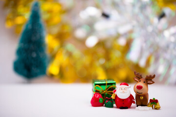 Santa Claus resin dolls stand alongside brown reindeer. The side are decorated with gift boxes, snow flakes and gold bells. Soft white floor. Faint bokeh on gold and silver backdrop and Christmas tree