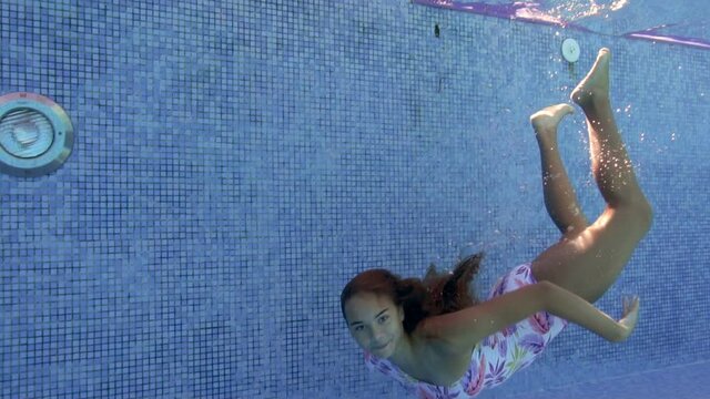 Cute, smiling teen girl dives and swims at the bottom of the pool in clear water. She poses for the camera with beautiful long hair and smiles. Portrait. Concept. Slow motion