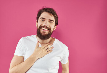 Happy guy in a white T-shirt on a pink background with a thick beard is gesturing with his hands
