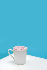 A white mug with marshmallows on a white table against a blue wall with copy space.