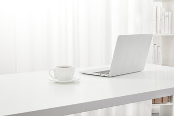 Closeup of a laptop and a cup of coffee on a white desk, next to a bookcase indoors