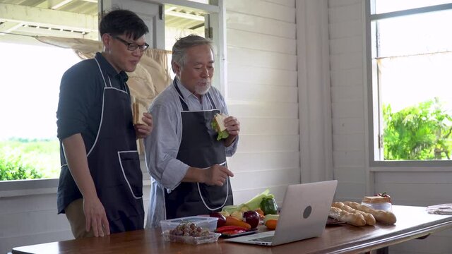 happy asian young son and senior old father cooking online class on laptop computer together making fresh vegetables food in kitchen at home