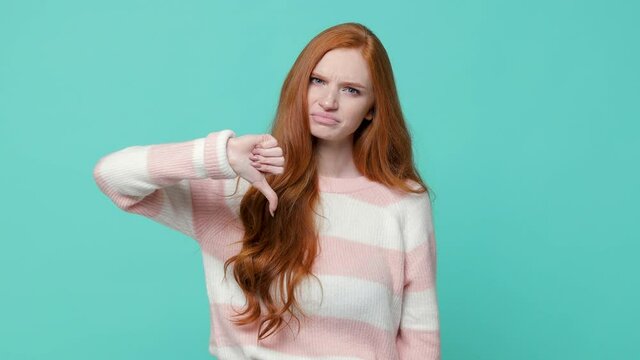 Dissatisfied ginger redhead young girl wearing white pink sweater posing isolated on blue turquoise color background in studio. People lifestyle concept. Say no with finger gesture showing thumb down