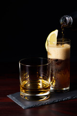 Whiskey neat and cola with ice and lemon wedge served on slate plate. Dark red wooden table, high resolution