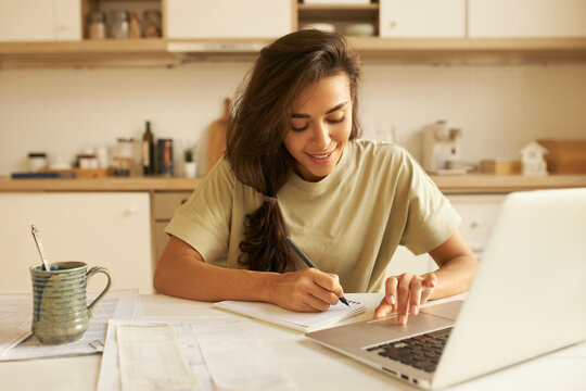 Indoor image of cute Arabic student girl in t-shirt sitting at dining table in front of open laptop, studying distantly, drinking morning coffee and making notes in copybook, listening to lecture